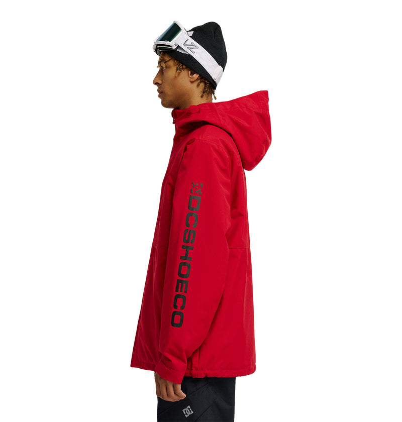 A person wearing a red DC Shoes Basis Jacket 10K with “DCSHOECO” on the sleeve, black pants, a black beanie, and white ski goggles stands in profile against a white background.