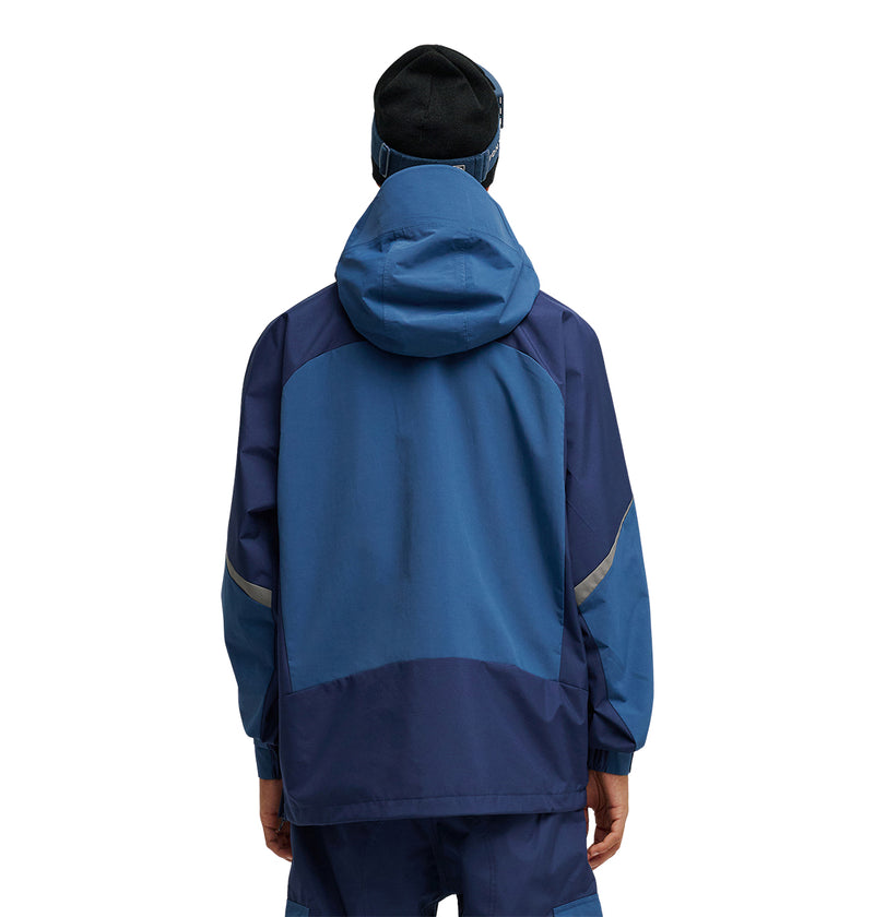 A person is seen from behind wearing the DC Shoes Mountain Lab Anorak 10K—a blue hooded jacket paired with matching pants, engineered for outstanding snow performance and breathable protection. They are also wearing a black beanie, and the background is plain white.