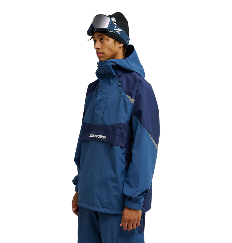 A person in a blue ski jacket and pants stands facing left, wearing ski goggles on a black beanie. The jacket, the Mountain Lab Anorak 10K by DC Shoes, is designed for exceptional snow performance and breathable protection.
