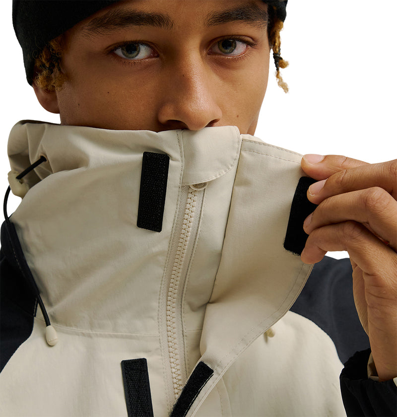 A person in a black beanie fastens the velcro collar of the DC Shoes Mountain Lab Anorak 10K snow jacket in beige and black, partially covering their face.