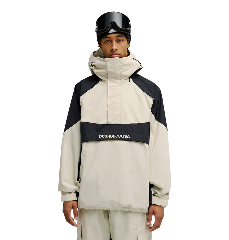 A person stands against a plain white background, ready for snow performance in the DC Shoes Mountain Lab Anorak 10K—a beige and black, hooded waterproof jacket featuring DCSHOECOUSA on the front—paired with beige pants and black ski goggles on their head, ensuring breathable protection.