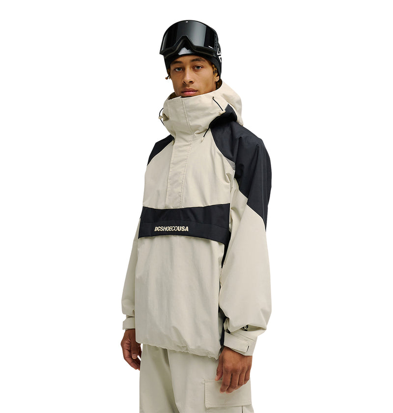 A person wearing the DC Shoes Mountain Lab Anorak 10K beige and black hooded snow jacket, paired with beige pants and black ski goggles, stands facing forward against a white background.
