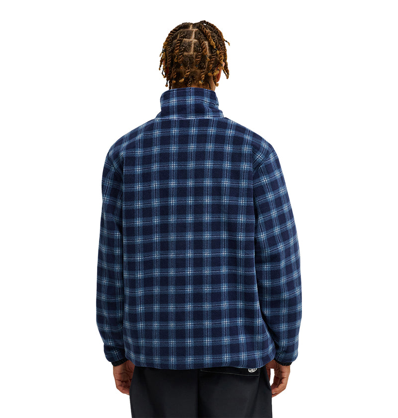 A person with braided hair is shown from the back, wearing the DC Shoes Park Reversible Anorak in blue and black plaid, paired with black pants, against a plain white background.