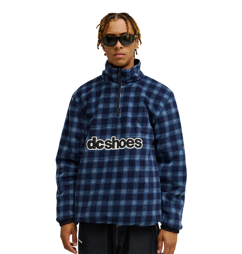 A person wearing the DC Shoes Park Reversible Anorak, featuring a blue plaid zip-up design with “dcshoes” on the front, stands against a white background and showcases its reversible style and advanced waterproofing for enhanced snow performance.