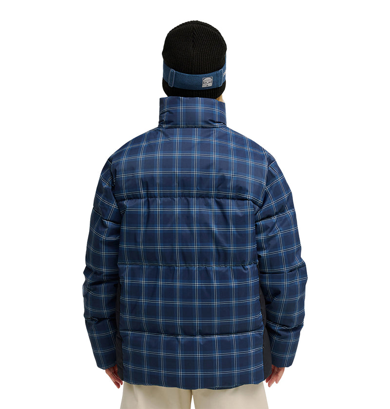 A person is seen from behind, wearing the DC Shoes Trackstar Puffer 10K jacket in blue plaid, paired with cream pants and a black beanie featuring a blue headband. The background is plain white.