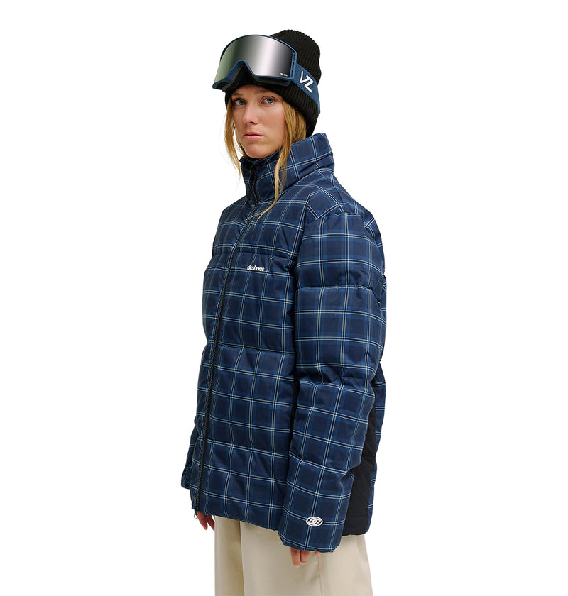 A woman wearing the DC Shoes Trackstar Puffer 10K blue plaid insulated winter jacket, beige pants, a black beanie, and ski goggles stands facing slightly to the side against a white background.