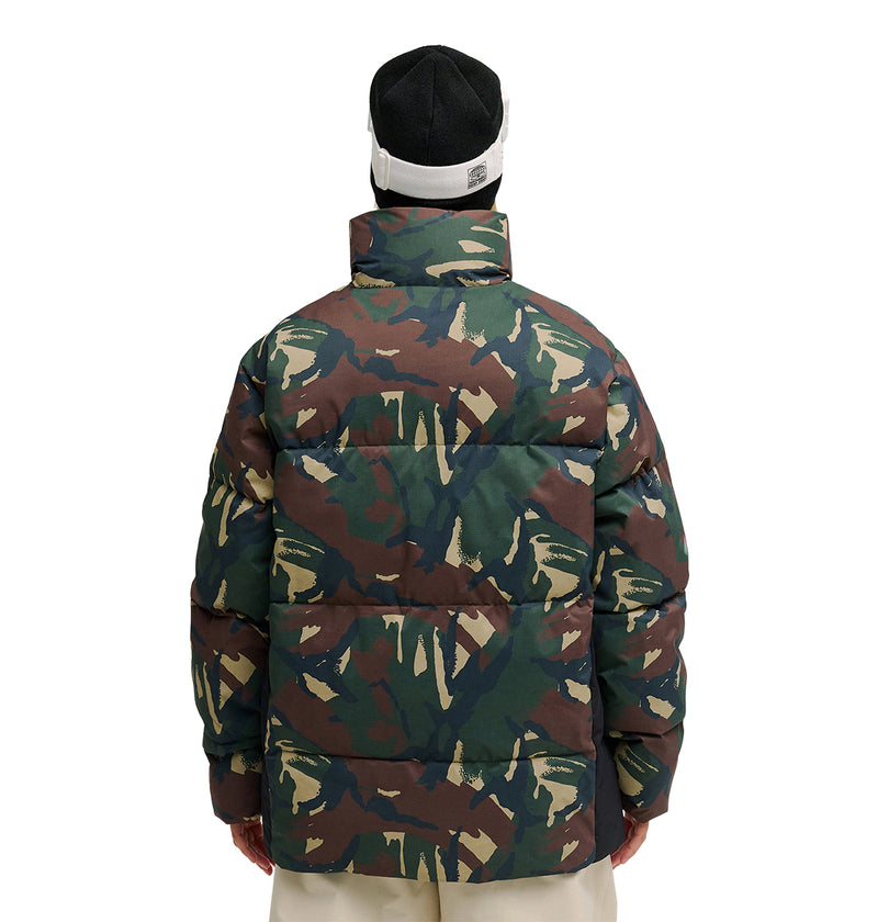 A person is shown from the back wearing the DC Shoes Trackstar Puffer 10K jacket in a green, brown, and beige camouflage print, paired with a black and white beanie, standing against a plain white background.