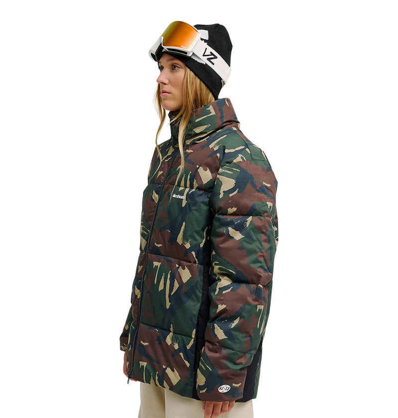 A woman stands in profile against a white background, wearing the DC Shoes Trackstar Puffer 10K insulated camouflage jacket, beige pants, and a black beanie with ski goggles.