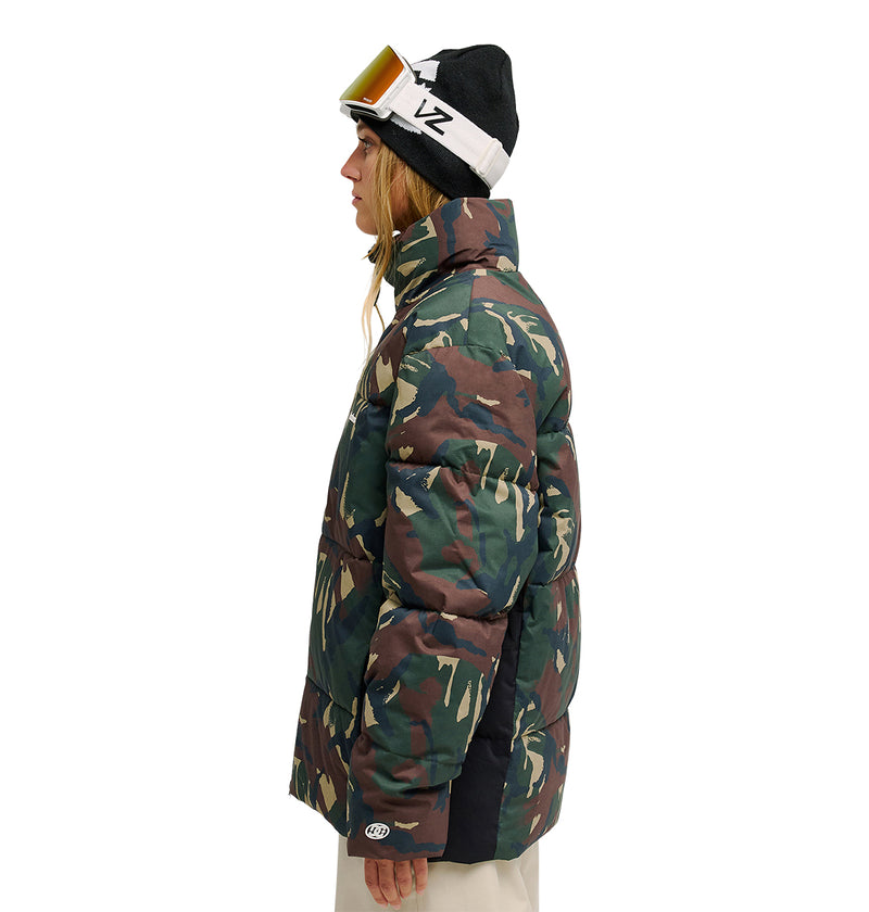 A person in profile wears the DC Shoes Trackstar Puffer 10K—an insulated camo-pattern jacket—paired with a black winter hat topped with white goggles, long hair visible, against a plain white background.