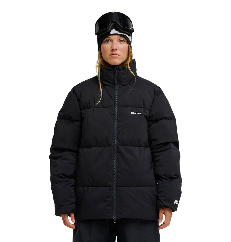 A person stands facing forward, wearing the DC Shoes Trackstar Puffer 10K jacket in black, which features breathable, waterproof protection. They have black ski goggles resting on their forehead and a black beanie. The jacket displays a small white DC Shoes logo on the left chest and left sleeve. The background is plain white.