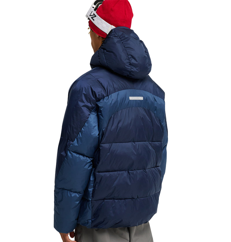 A person wearing a red beanie and a dark blue DC Shoes Powder Puffer 10K jacket with a hood stands facing away from the camera. The jacket, featuring a shiny finish and horizontal stitching, is designed for excellent insulation and snow performance.