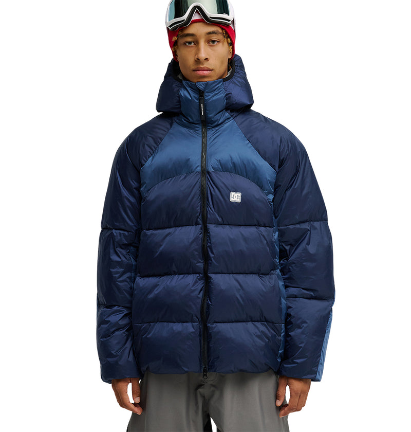 A person stands facing the camera against a white background, wearing the DC Shoes Powder Puffer 10K blue jacket, renowned for its superior insulation and waterproofing, along with gray pants and ski goggles on their head.