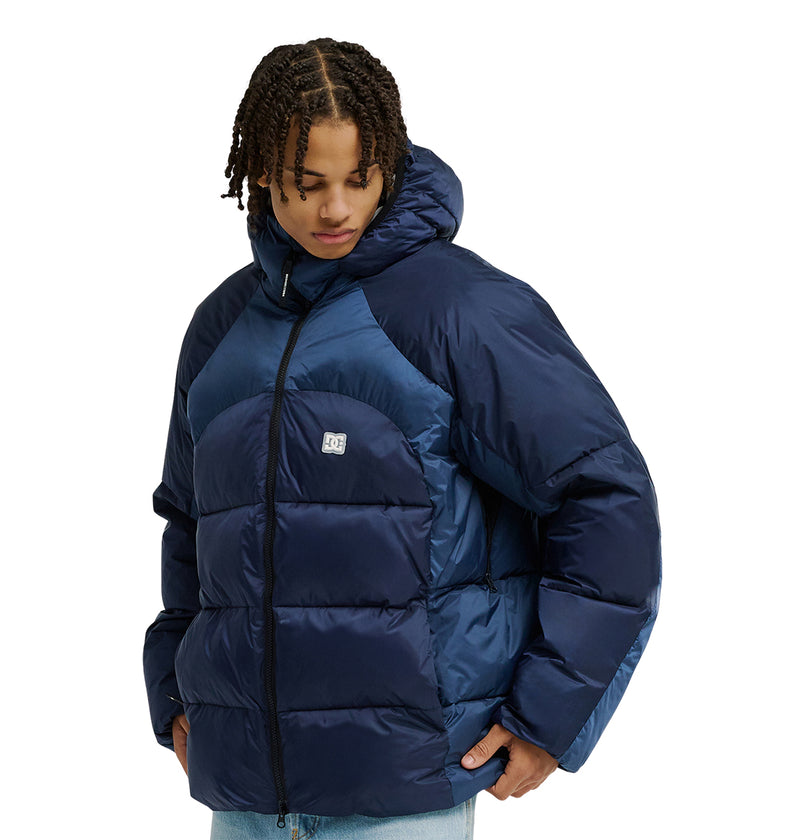 A person with short, twisted hair wears the DC Shoes Powder Puffer 10K jacket—a shiny navy blue puffer with a hood. Engineered for top snow performance and superior insulation, this jacket features a front zipper and a small DC Shoes logo on the chest. The individual is looking down and to the side.