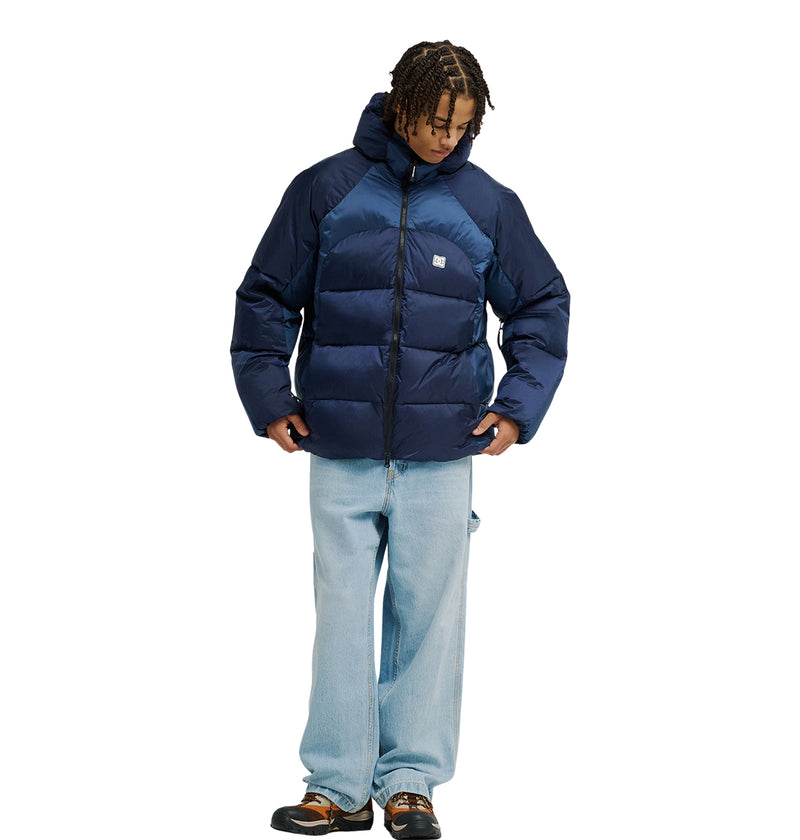 A person wearing the DC Shoes Powder Puffer 10K, a navy blue hooded jacket known for excellent insulation and waterproofing to enhance snow performance, paired with light blue baggy jeans and brown shoes, is looking down and slightly adjusting the jacket. The background is plain white.