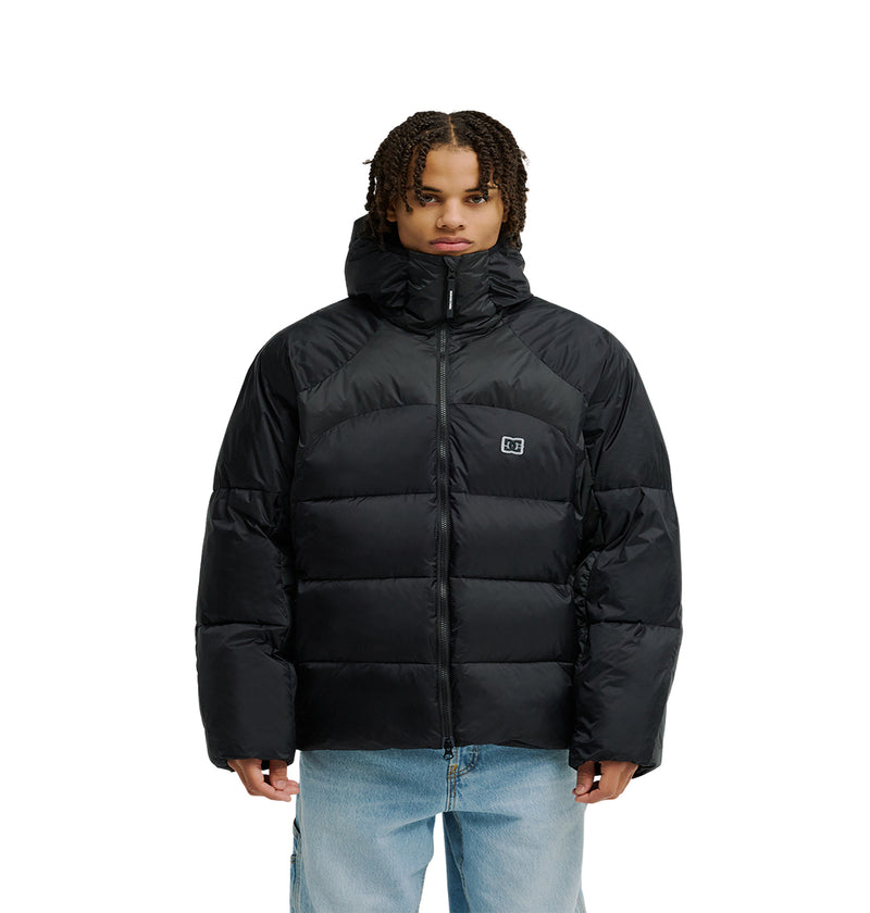 A person stands facing forward, wearing the DC Shoes Powder Puffer 10K—a black puffer jacket with a hood designed for breathable protection. The jacket features a small square logo on the chest and is zipped up. The individual has short, curly hair and is also wearing light blue jeans.