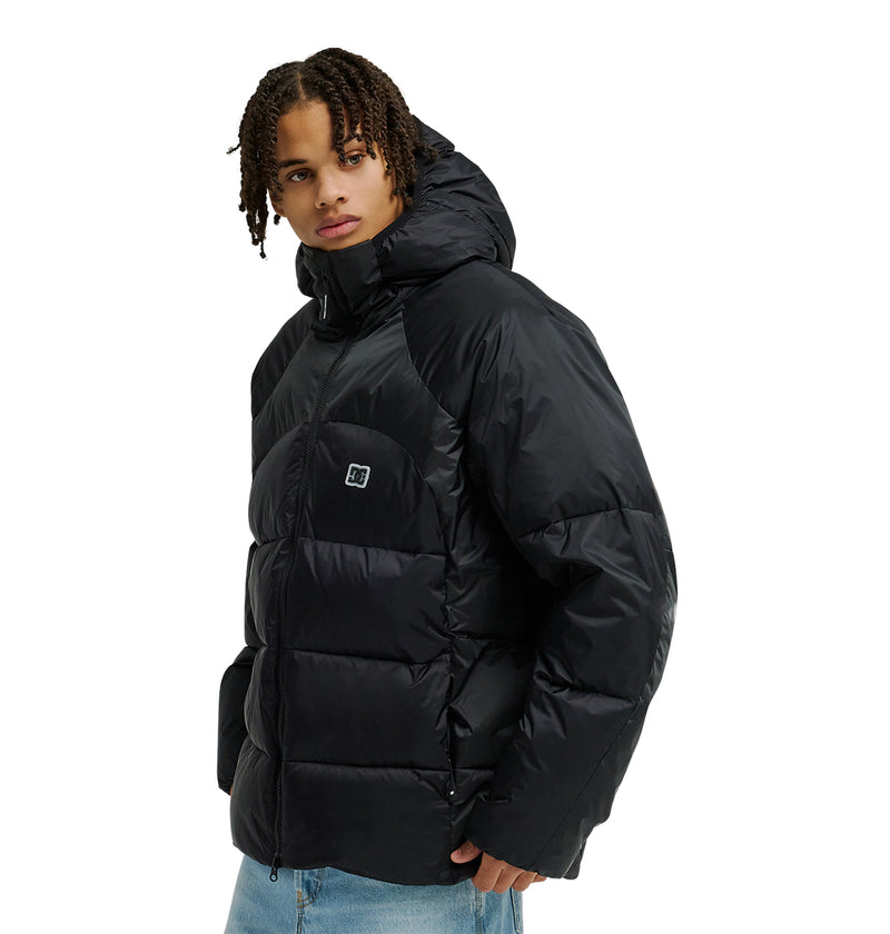 A person wearing the DC Shoes Powder Puffer 10K, a black hooded jacket engineered for breathable protection and snow performance, paired with light blue jeans, stands facing slightly to the side while looking toward the camera.