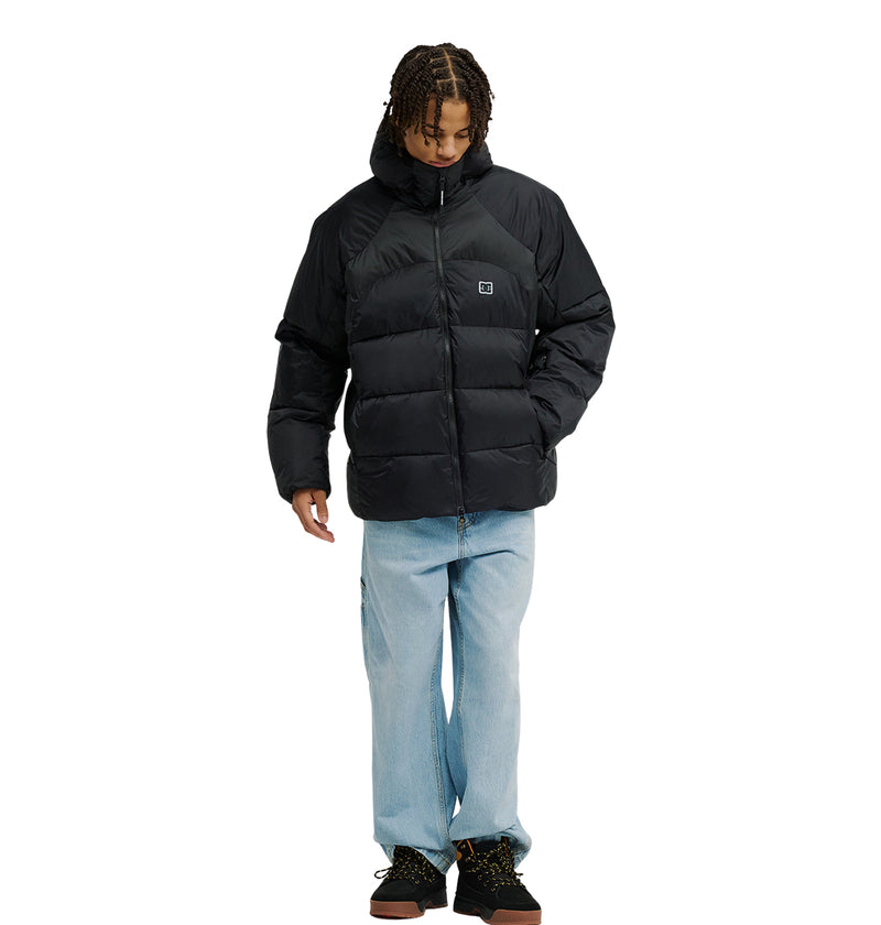 A person stands facing forward against a plain white background, looking down with hands in their pockets, dressed in the DC Shoes Powder Puffer 10K jacket—featuring breathable protection—paired with light blue jeans and dark shoes.