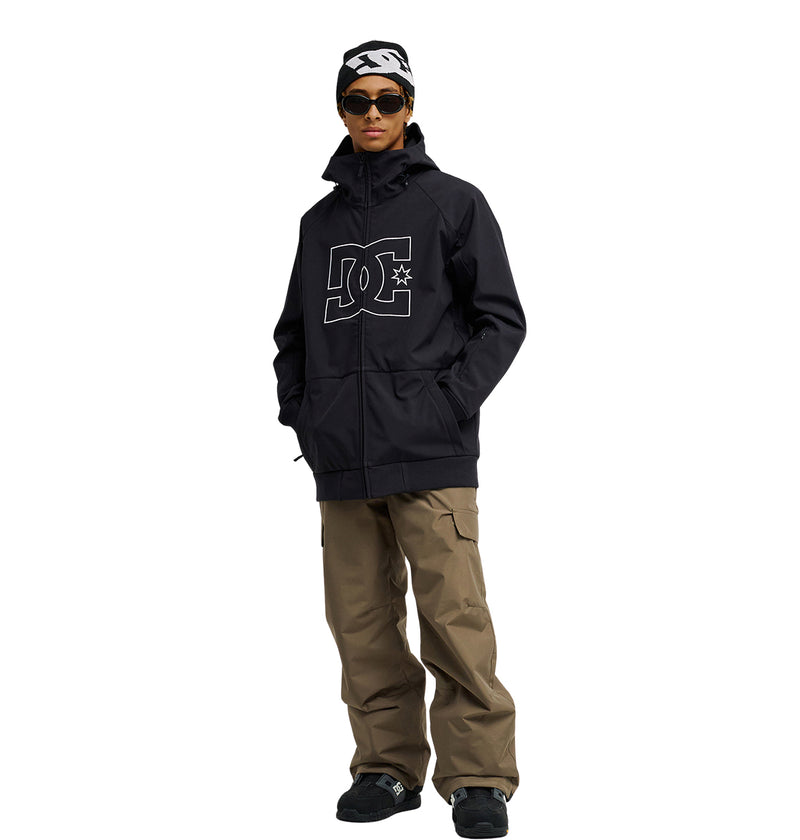 A person stands with hands in pockets against a white background, wearing the DC Shoes Spectrum Softshell Jacket—a black style featuring a large DC logo—paired with khaki cargo pants, black shoes, dark sunglasses, and a black-and-white beanie. The Spectrum Softshell Jacket offers breathable protection for comfort and style in any setting.