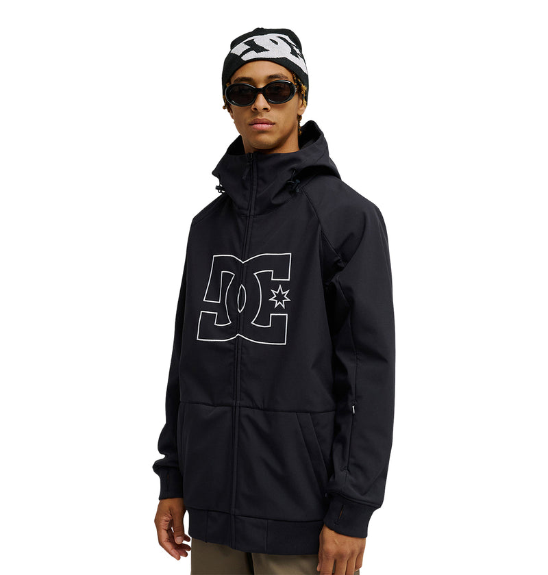A person stands facing forward against a white background, showcasing the DC Shoes Spectrum Softshell Jacket—a black jacket with a prominent DC logo on the front—paired with sunglasses, a patterned black-and-white beanie, and light-colored pants for breathable protection and everyday comfort.