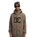 A person wearing sunglasses, a black beanie, and the DC Shoes Spectrum Softshell Jacket featuring a large DC logo on the front stands facing the camera against a white background.