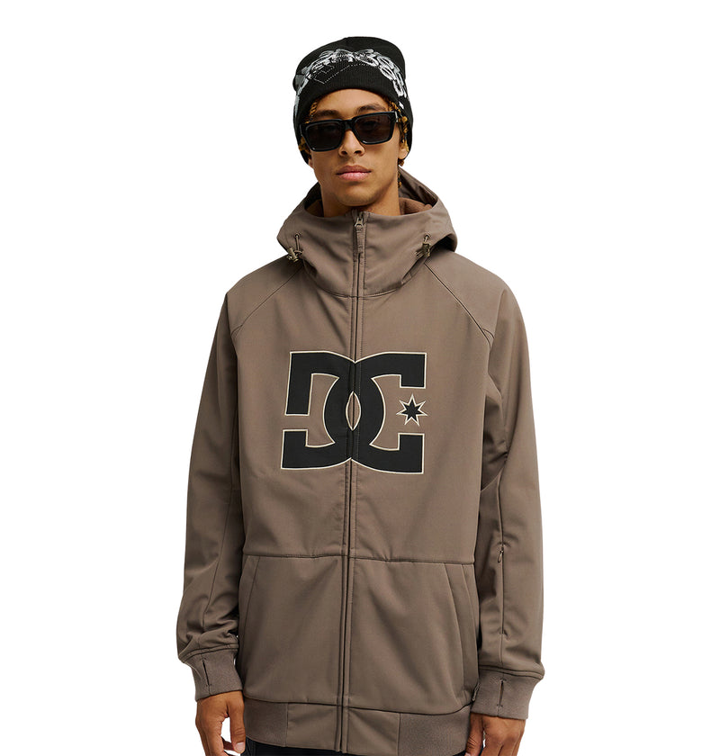 A person wearing sunglasses, a black beanie, and the DC Shoes Spectrum Softshell Jacket featuring a large DC logo on the front stands facing the camera against a white background.