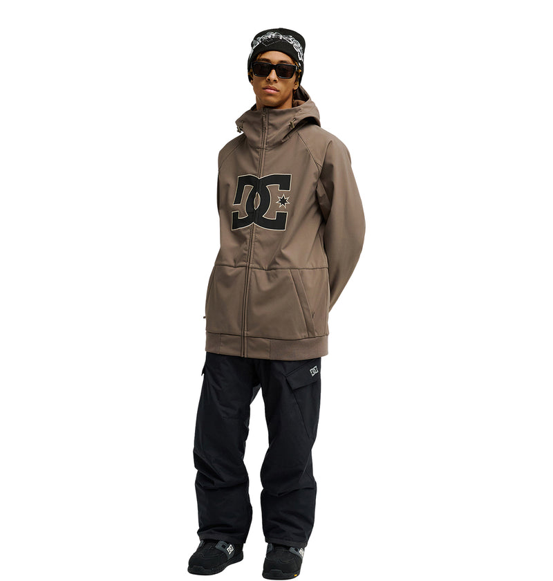 A person stands against a plain white background wearing the DC Shoes Spectrum Softshell Jacket in tan, paired with black pants, black sneakers, sunglasses, and a patterned headband—a look designed for snow performance yet styled for any weather.