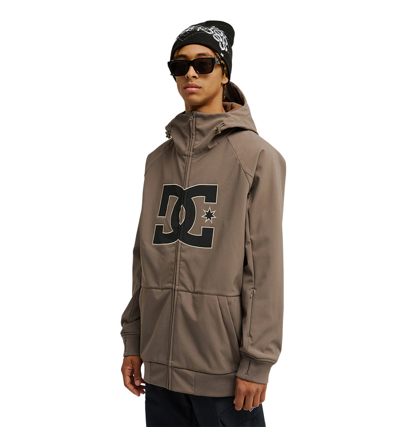 A person wearing sunglasses, a black beanie, and the DC Shoes Spectrum Softshell Jacket featuring a large DC logo on the front stands against a white background.