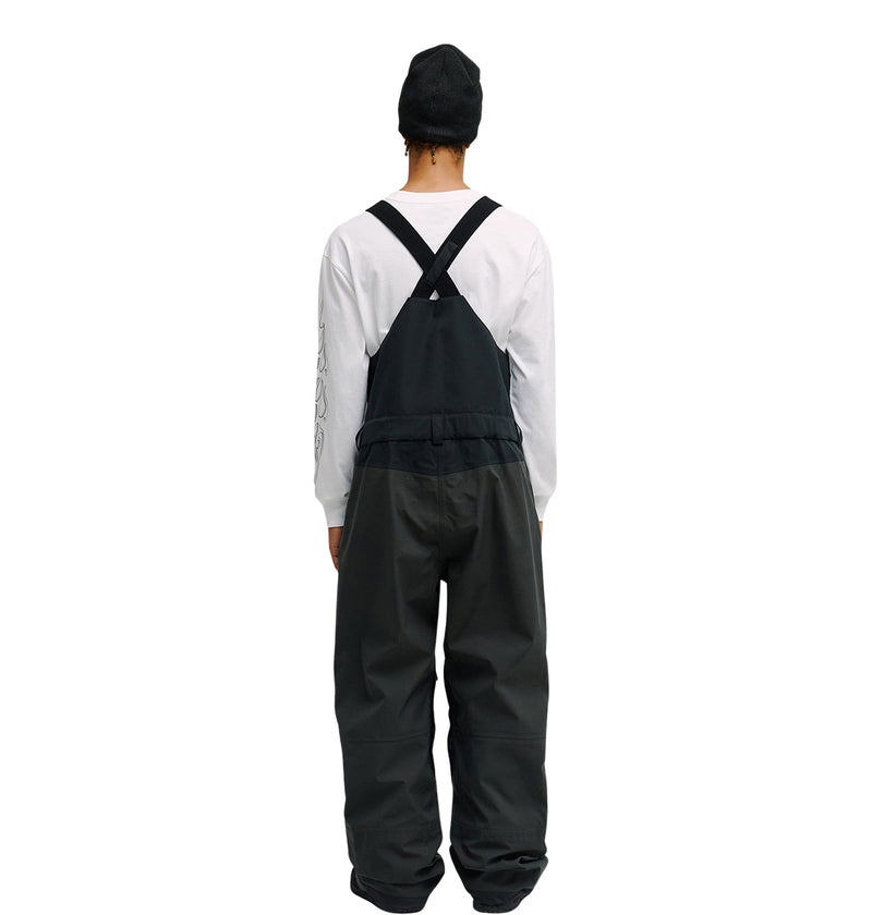 A person wearing a black beanie, a white long-sleeve shirt, and the DC Shoes Storm Division 3L Bib 30K black overalls with breathable protection is standing facing away from the camera against a white background.