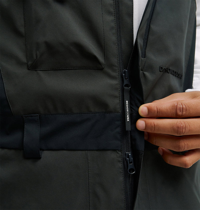 A person’s hand is shown zipping up the Storm Division 3L Bib 30K by DC Shoes, a dark-colored jacket featuring a chest pocket and a visible DC Shoes brand label near the zipper. This stylish outerwear delivers breathable protection, making it perfect for active days outdoors.