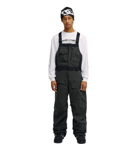 A person stands facing forward with hands at their sides against a plain white background, wearing the DC Shoes Storm Division 3L Bib 30K over dark attire, a white long-sleeve shirt with text, and a black beanie—showcasing this bib’s breathable protective design.
