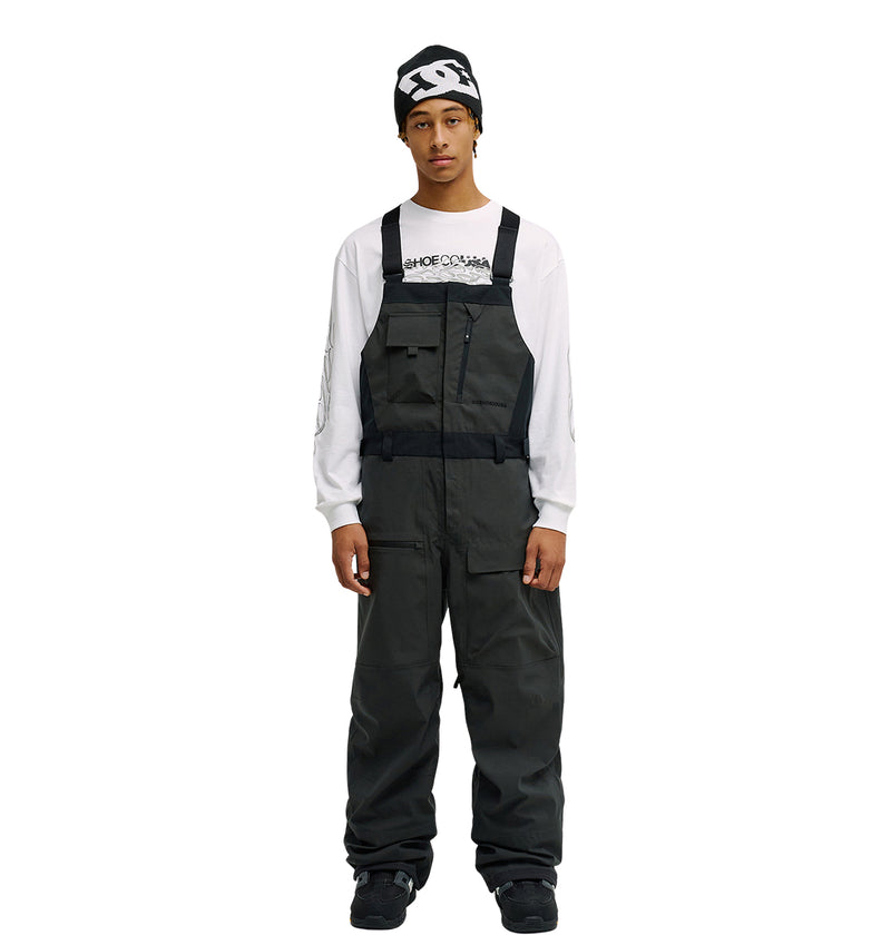 A person stands facing forward with hands at their sides against a plain white background, wearing the DC Shoes Storm Division 3L Bib 30K over dark attire, a white long-sleeve shirt with text, and a black beanie—showcasing this bib’s breathable protective design.