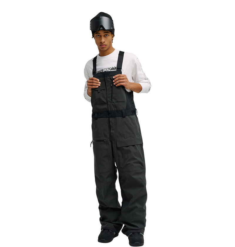 A person is wearing a white long-sleeve shirt, black Storm Division 3L Bib 30K pants by DC Shoes—engineered for breathable protection—along with black shoes and ski goggles on their head, standing against a white background.