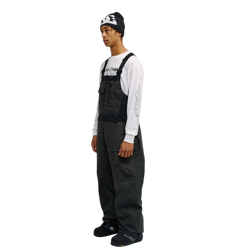 A person stands sideways against a white background wearing the DC Shoes Storm Division 3L Bib 30K in black, paired with a black beanie, a white graphic long-sleeve shirt, and black shoes—showcasing breathable protection engineered for enhanced snow performance.