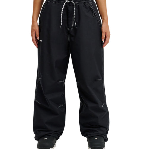 A person is shown from the waist down, standing against a white background, wearing loose-fitting DC Shoes Trackstar Pants 10K in black with white stitching, a drawstring waist, cargo pockets, and paired with black shoes.
