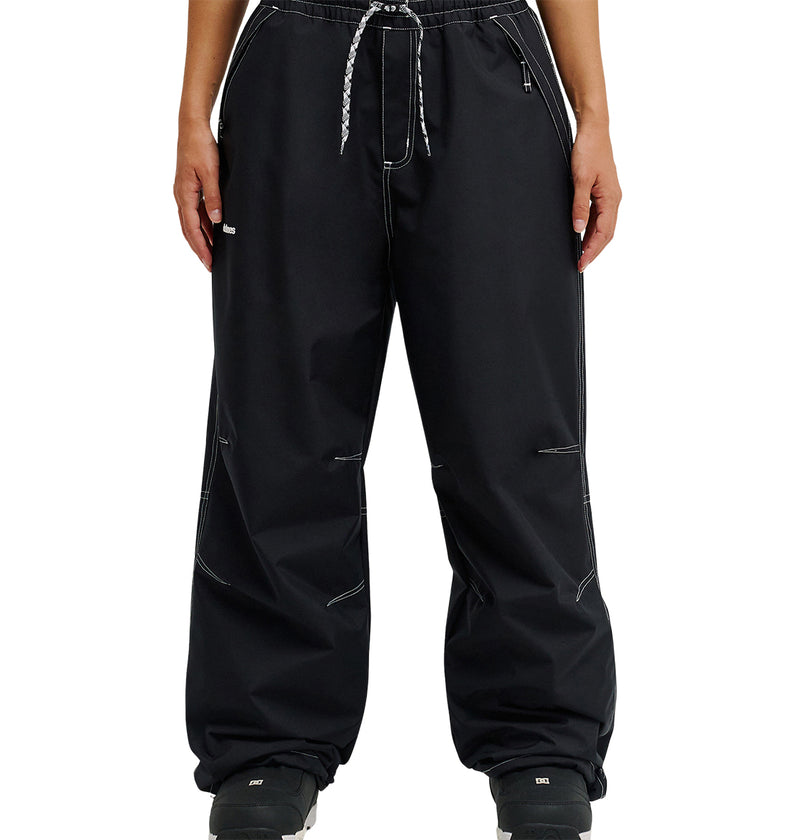 A person is shown from the waist down, standing against a white background, wearing loose-fitting DC Shoes Trackstar Pants 10K in black with white stitching, a drawstring waist, cargo pockets, and paired with black shoes.