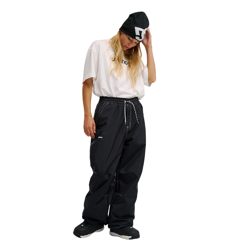 A person with long hair, wearing a black beanie, a white t-shirt, and baggy DC Shoes Trackstar Pants 10K, stands looking down while adjusting their hat. Their casual and sporty outfit subtly nods to snow performance with the enhanced waterproofing of the Trackstar Pants 10K.