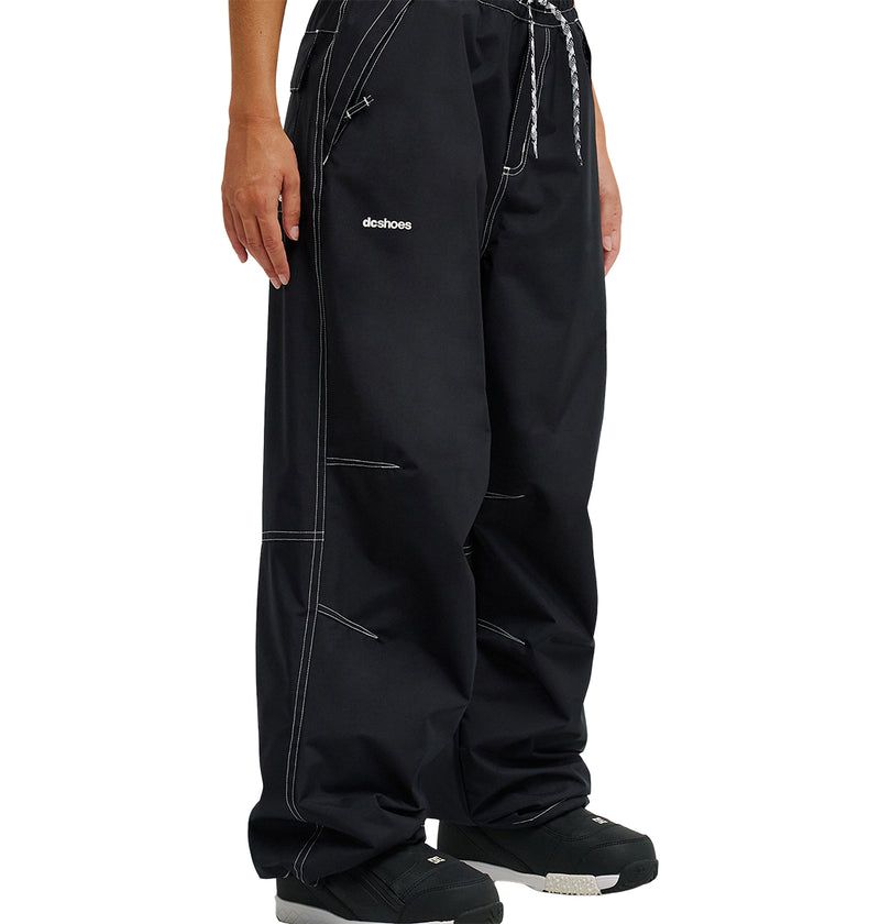 A person is seen from the waist down wearing loose-fitting black Trackstar Pants 10K by DC Shoes, featuring white stitching, a black-and-white drawstring, and “DC Shoes” printed on the left thigh. The pants are designed for enhanced snow performance and paired with black shoes.