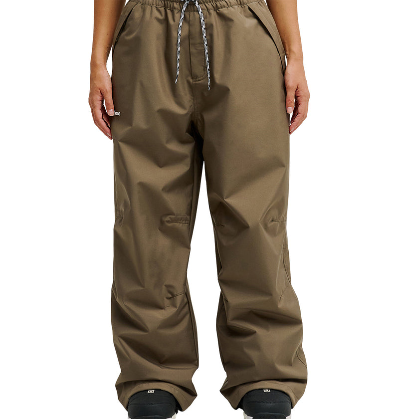 A person wearing DC Shoes Trackstar Pants 10K—loose-fitting brown waterproof pants with a drawstring waist—paired with black shoes, photographed from the waist down against a plain white background. These pants are an ideal choice for breathable protection in all conditions.