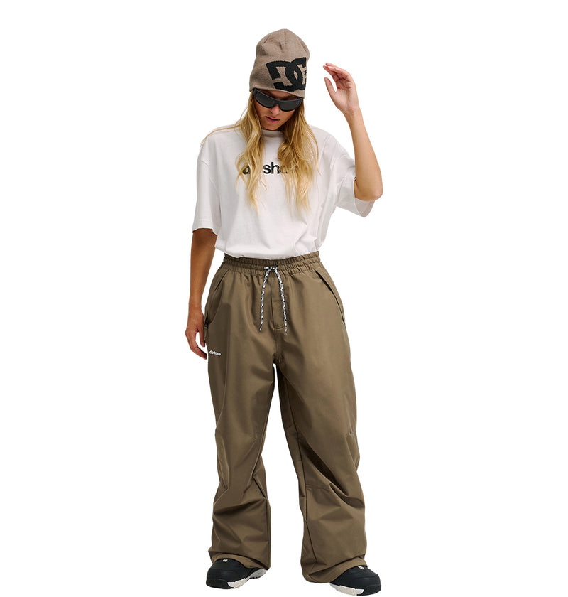 A person wearing a beige beanie, black sunglasses, a white t-shirt, and DC Shoes Trackstar Pants 10K in brown with black shoes stands against a white background.