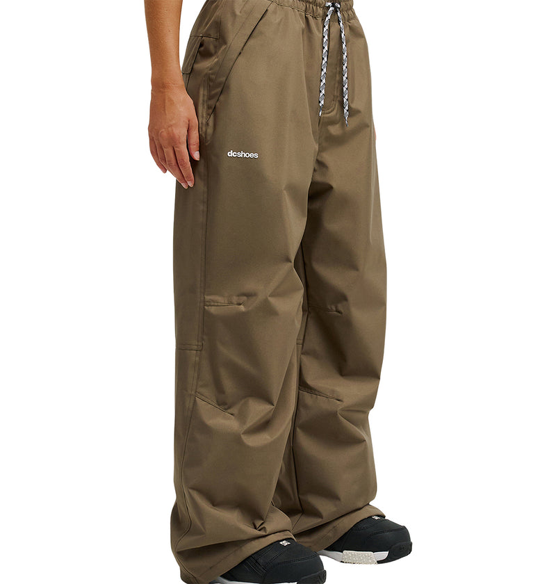 A person wearing the DC Shoes Trackstar Pants 10K—loose-fitting brown waterproof pants with a black and white drawstring, black shoes, and a visible DC Shoes logo on the left thigh—stands against a plain white background; these pants are designed for breathable protection.