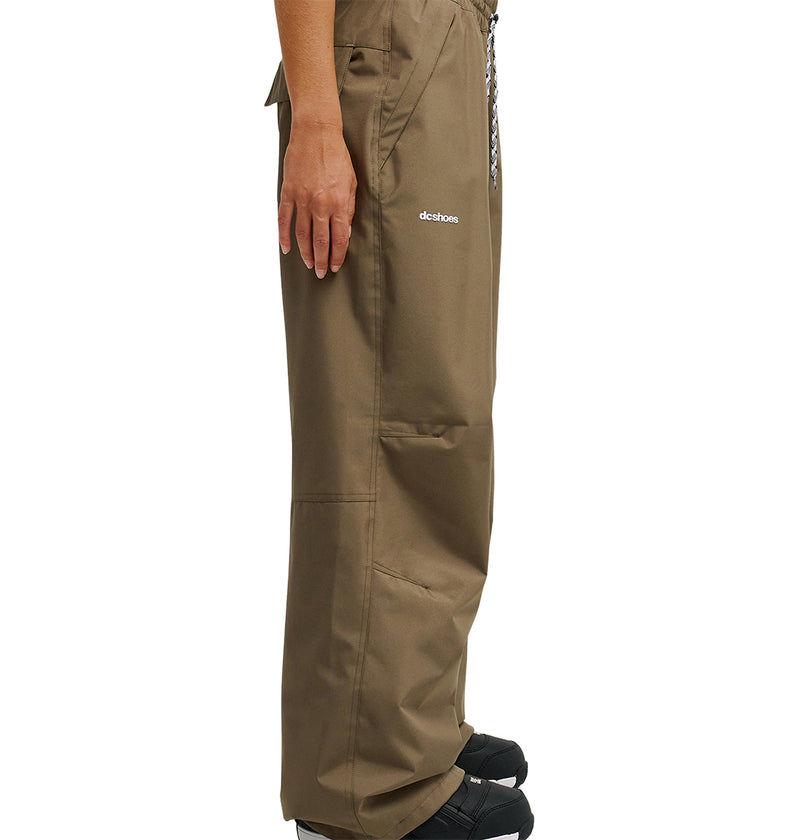A person stands sideways wearing loose brown DC Shoes Trackstar Pants 10K with pockets and black shoes. The pants, featuring breathable waterproof protection, have the word “detchos” printed near the pocket. Only the lower half of the body is visible.