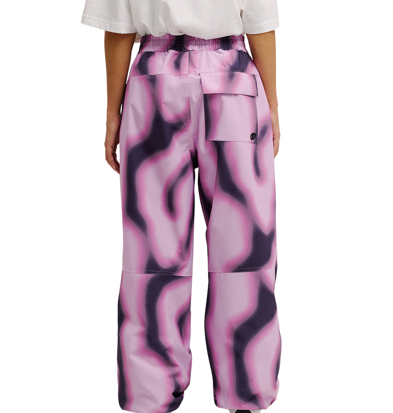 A person is seen from the back wearing DC Shoes Trackstar Pants 10K—loose, high-waisted waterproof pants with a pink and black wavy pattern. The pants provide breathable protection, feature a large right-side pocket, and have elastic at the waist and ankles.