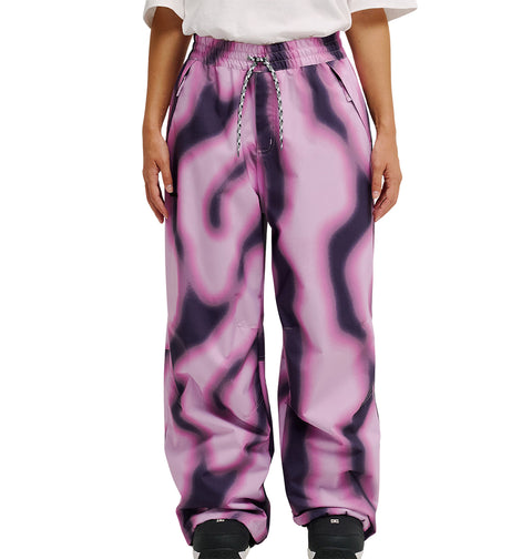 A person wearing DC Shoes Trackstar Pants 10K—loose, high-waisted, pink waterproof pants with a black wavy pattern—along with a white shirt and black shoes, standing against a plain white background.