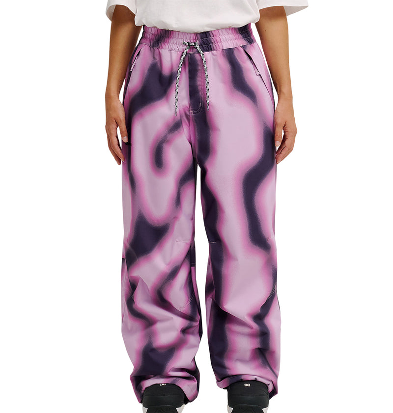 A person wearing DC Shoes Trackstar Pants 10K—loose, high-waisted, pink waterproof pants with a black wavy pattern—along with a white shirt and black shoes, standing against a plain white background.