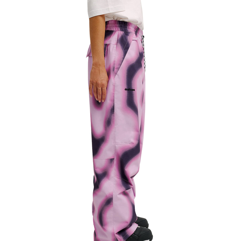 A person is shown in side profile against a white background, wearing the DC Shoes Trackstar Pants 10K featuring a pink and black abstract pattern and paired with a white shirt.
