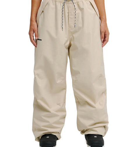 A person is wearing the DC Shoes Trackstar Pants 10K—loose-fitting beige pants with an elastic drawstring waist, breathable protection, and a black logo on the left thigh—paired with black shoes and shown against a plain white background.