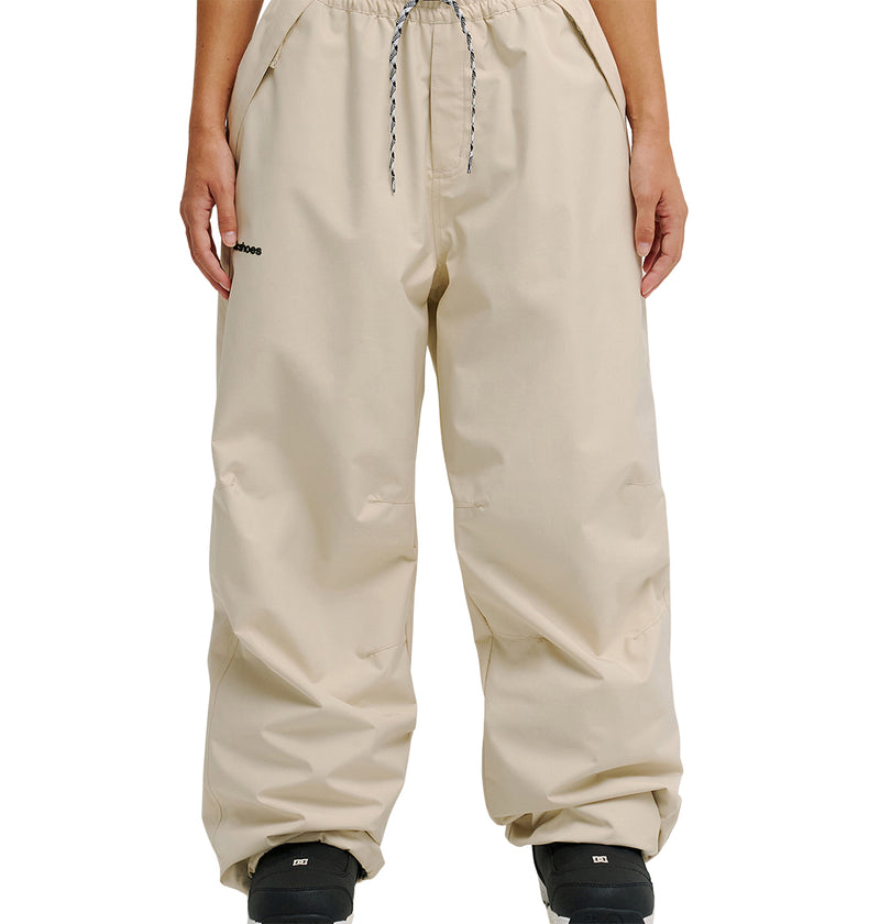 A person is wearing the DC Shoes Trackstar Pants 10K—loose-fitting beige pants with an elastic drawstring waist, breathable protection, and a black logo on the left thigh—paired with black shoes and shown against a plain white background.
