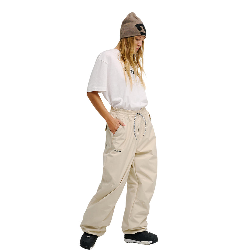 A person wearing a tan beanie, oversized white t-shirt, DC Shoes Trackstar Pants 10K in beige—designed for breathable protection—and black shoes stands against a white background, looking down with hands in pockets.