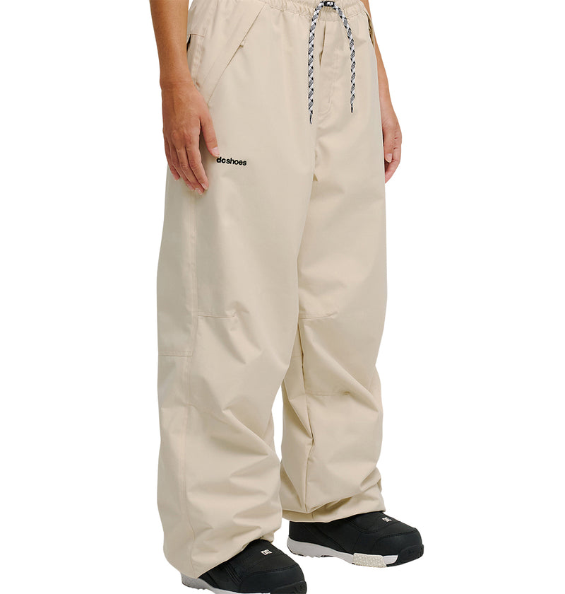 A person wearing loose beige DC Shoes Trackstar Pants 10K with a drawstring waist, black boots, and a visible logo on the left leg, shown from the waist down.