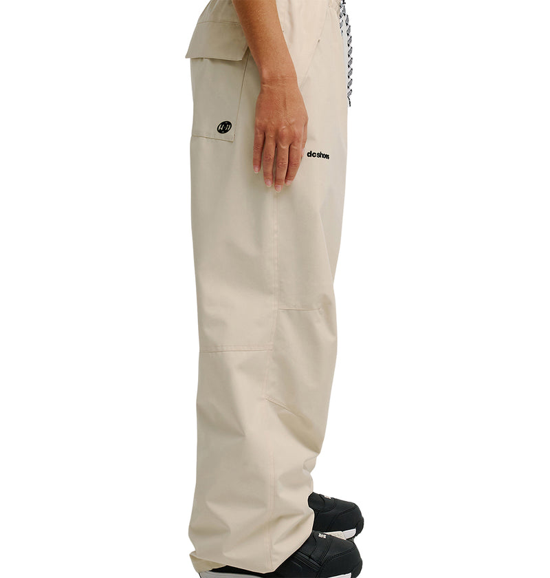 A person standing sideways, with only their lower body visible against a white background, is wearing the DC Shoes Trackstar Pants 10K in beige featuring small black logos and paired with black shoes. These pants provide breathable protection for enhanced comfort.